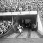 Heinäkuun 19. päivä vuonna 1952. Kello on 14.28. Paavo Nurmi tuo olympiastadionille olympiasoihdun. Eturivissä Raimo Mykkänen tuulettaa hetkeä valokuvaajan oikealla puolella valkoisessa takissaan toinen käsi pystyssä, toinen kaiteessa, hattu päässään. Seuraavana iltana Mykkänen menee yöunille stadionin läheisen pensaikon alle, jotta rahat riittävät kisojen seuraamiseen ja kunnon majoitukseen vielä seuraavina päivinä. (Kuva: Suomen Urheilumuseo)