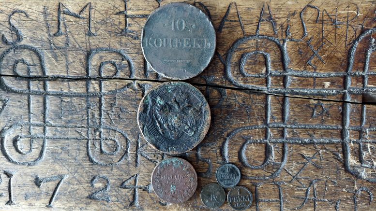 Kolmen kuninkaan uhrirahat” kuvattuna taikamerkein varustetun laudan päällä. Mukana lisäksi Aleksanteri II:n aikaiset pennit löytöpaikkana talon eteläisen nurkkakiven vierusta.