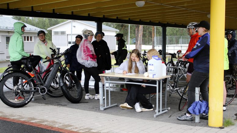 Ennen liikkeelle lähtöä olivat pyöräilijöiden hymyt vielä herkässä, vaikka sadevarusteet olivat tarpeen. Ilmoittautumispistekin piti siirtää koulun edustalle katoksen suojiin, jotta ilmoittautumisia pöydän ääressä keränneiden Reetta Aholan (vasemmalla) ja Kerttu Kynkäänniemen paperit pysyivät kuivina. Oikealla seisomassa Anne Kynkäänniemi. Kuvat: Ritva Piri
