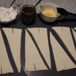 Croissant-torttujen valmistus alkaa. Ota esille tarpeet, kauli sulaneet taikinalevyt vähän pidemmiksi. Leikkaa levyt halki kolmioiksi ja tee pienet viillot.