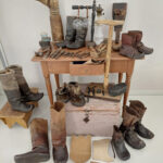 Pöydälle on asettunut suutarin koko työarsenaali. Pääosa työvälineistä on Koistis-Matin työkaluarkusta, joka on pöydän edustalla. Työhevonen seisoo arkun päällä, pieksujen kaavat löytyvät lattialta. Pöydällä suutarin vasara, veitset, riflauspyörät, naskalit, syrjäfummelit, pryylit, näverit, naula-, neula-, rasva- ja pikirasiat sekä lestin vetokoukut. Pieksuperhe on asettunut somasti myös potrettiin. Suutarin silmät kiiluvat sorvatussa. Kuva: Eino Mikkonen